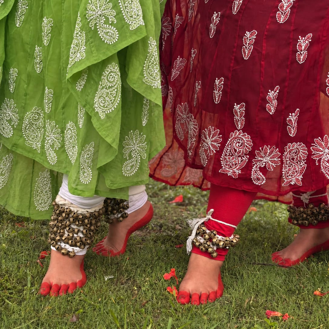 Close-up of two people standing on grass outdoors, wearing patterned green and red draped garments with white floral embroidery and red toe covers, along with ankle jewelry.
